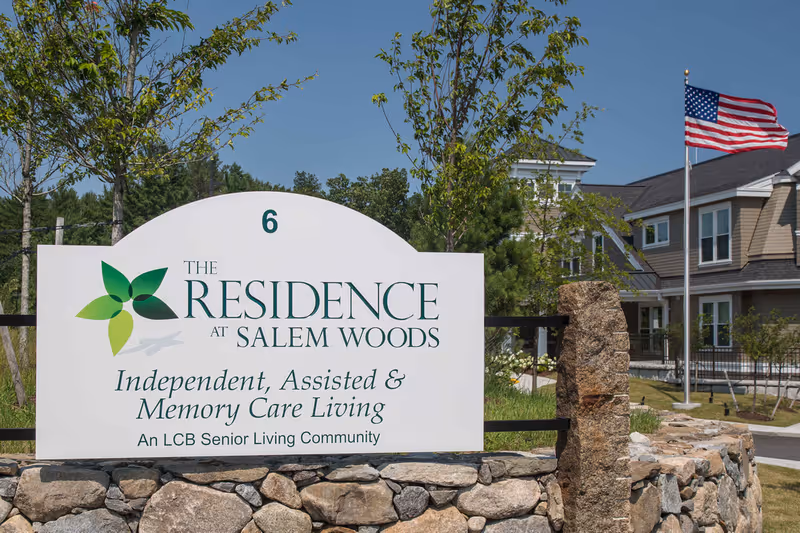 Outdoor view of a sign for The Residence at Salem Woods, an independent, assisted, and memory care senior living community. The sign is mounted on a stone base with trees and a building in the background, along with an American flag on a flagpole.