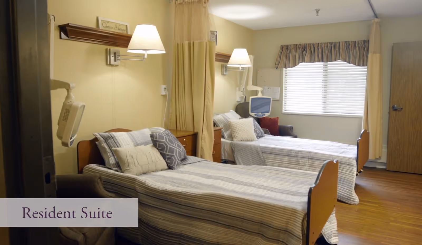 A resident suite in a senior living facility featuring two single beds with striped bedding and multiple pillows. Each bed has a wall-mounted lamp above it and a medical monitor attached. There is a window with blinds and a valance, a wooden door, and a curtain divider between the beds. The room has wooden flooring and neutral-colored walls.