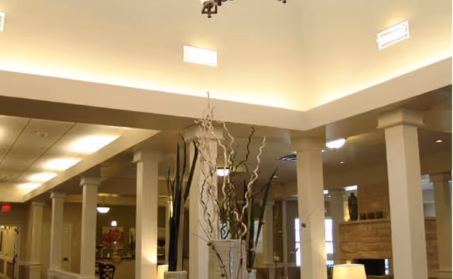 Interior view of a senior living facility common area with tall white columns, a high ceiling with recessed lighting, decorative plants in vases, and a stone fireplace in the background.