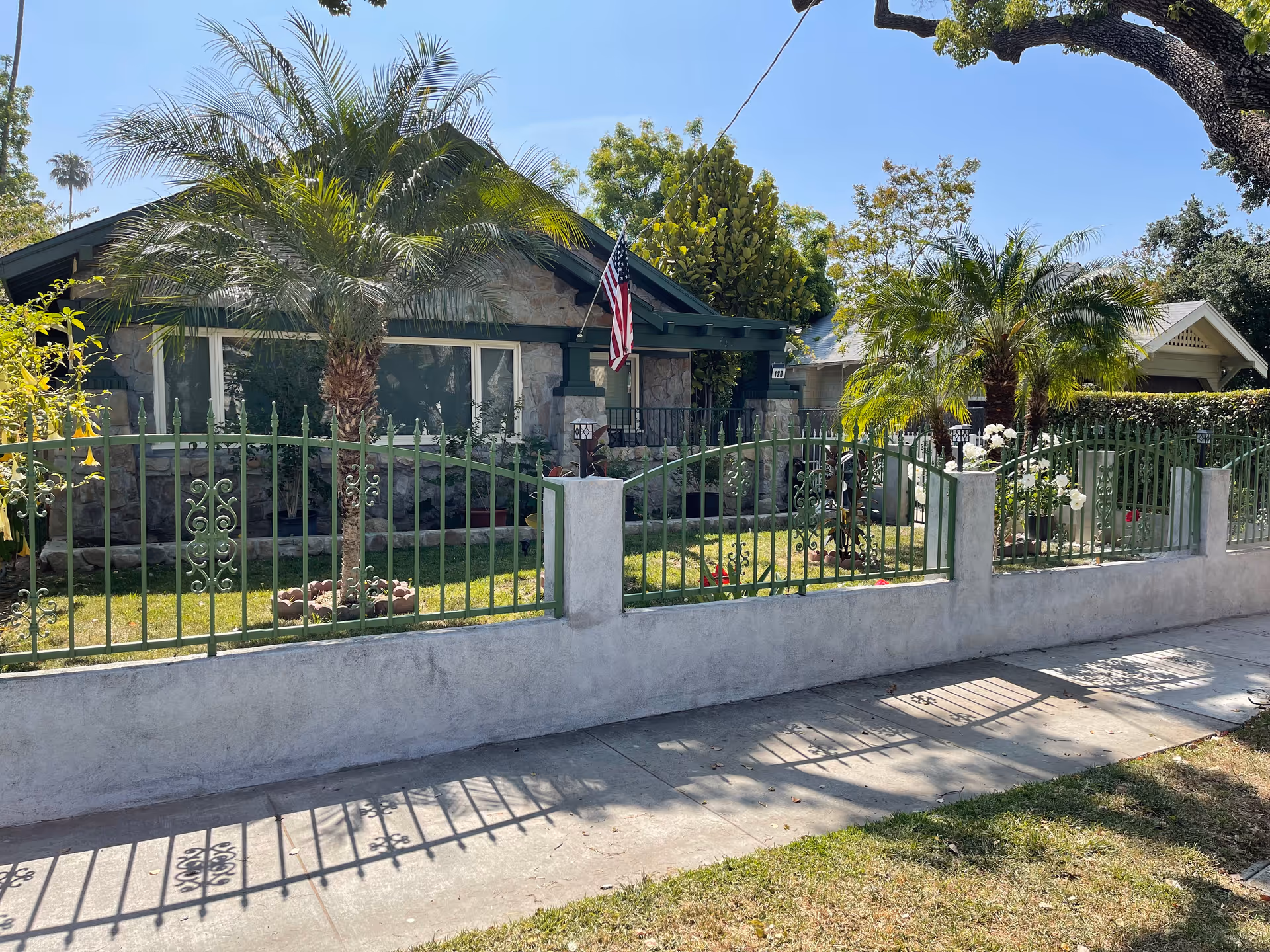 Single-story stone house with a green metal fence, palm trees, and an American flag in the front yard.