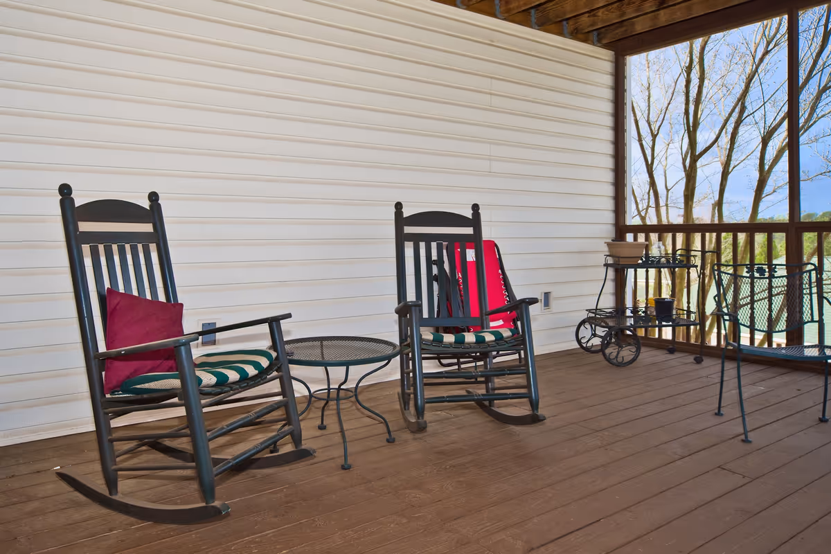 A covered wooden porch with two black rocking chairs with cushions and pillows, a small round metal table between them, and a metal plant stand with pots. The porch has a railing and overlooks trees and a clear blue sky.