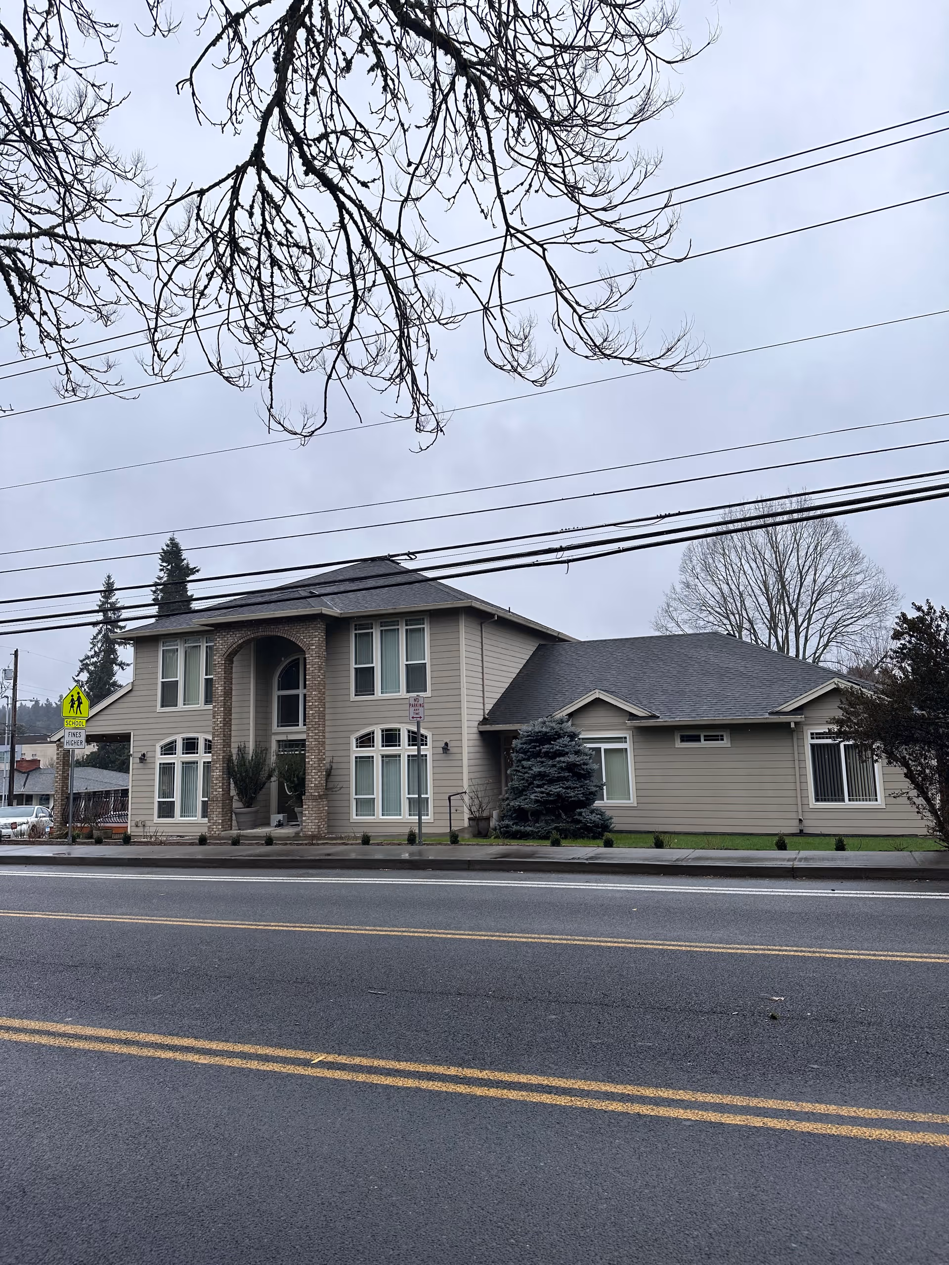 Exterior view of a two-story building with beige siding and a dark roof, featuring a central entrance with brick columns and large windows. The building is situated along a street with a pedestrian crossing sign visible. Leafless tree branches are seen overhead against a cloudy sky.