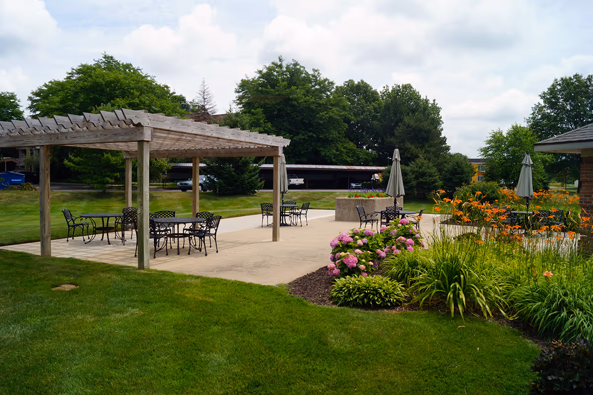 Outdoor patio area with metal tables and chairs under a wooden pergola and umbrellas, surrounded by green grass, flowering plants, and trees under a partly cloudy sky.