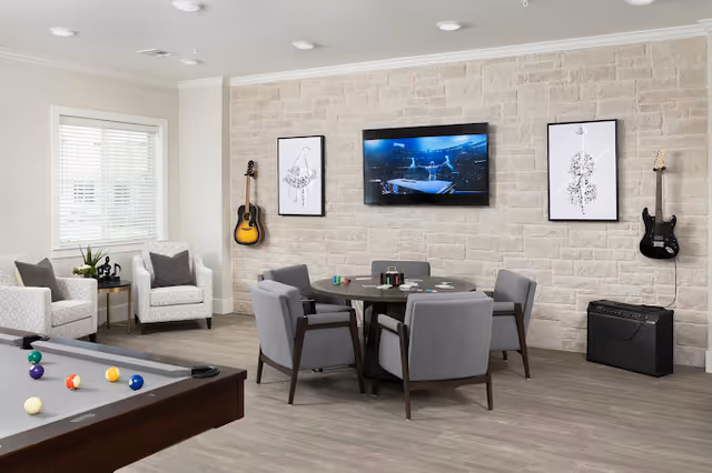 A modern recreational room with a pool table in the foreground, a round table with four gray chairs in the center, and two guitars mounted on a light stone wall. A flat-screen TV is mounted on the wall between two framed musical instrument artworks. Two white armchairs with gray cushions and a small side table with a plant are near a window with white blinds.