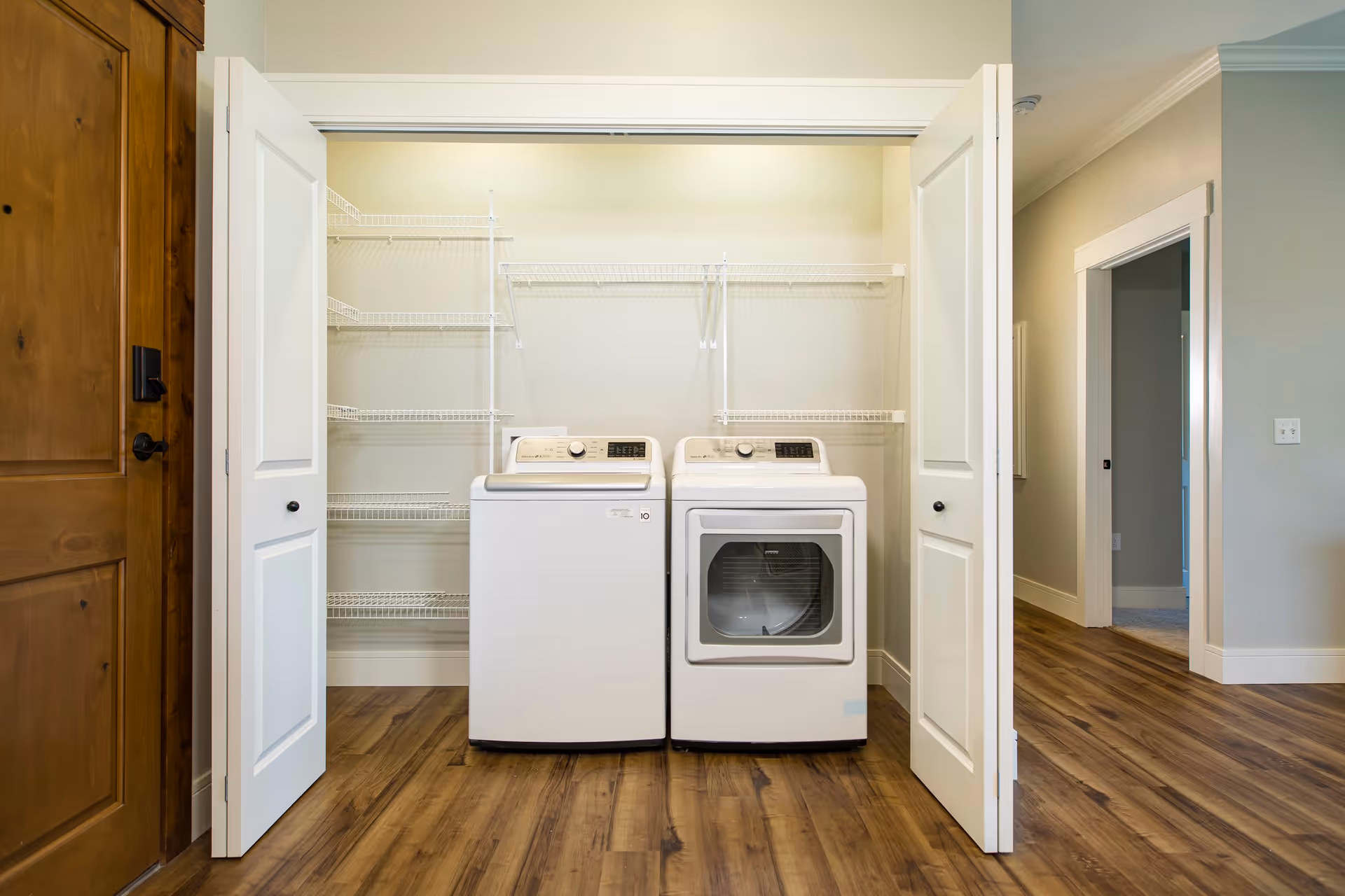 Laundry area with a white washing machine and dryer side by side inside a closet with open white double doors. The closet has multiple white wire shelves on the walls. The floor is wood and a wooden door is visible on the left side.
