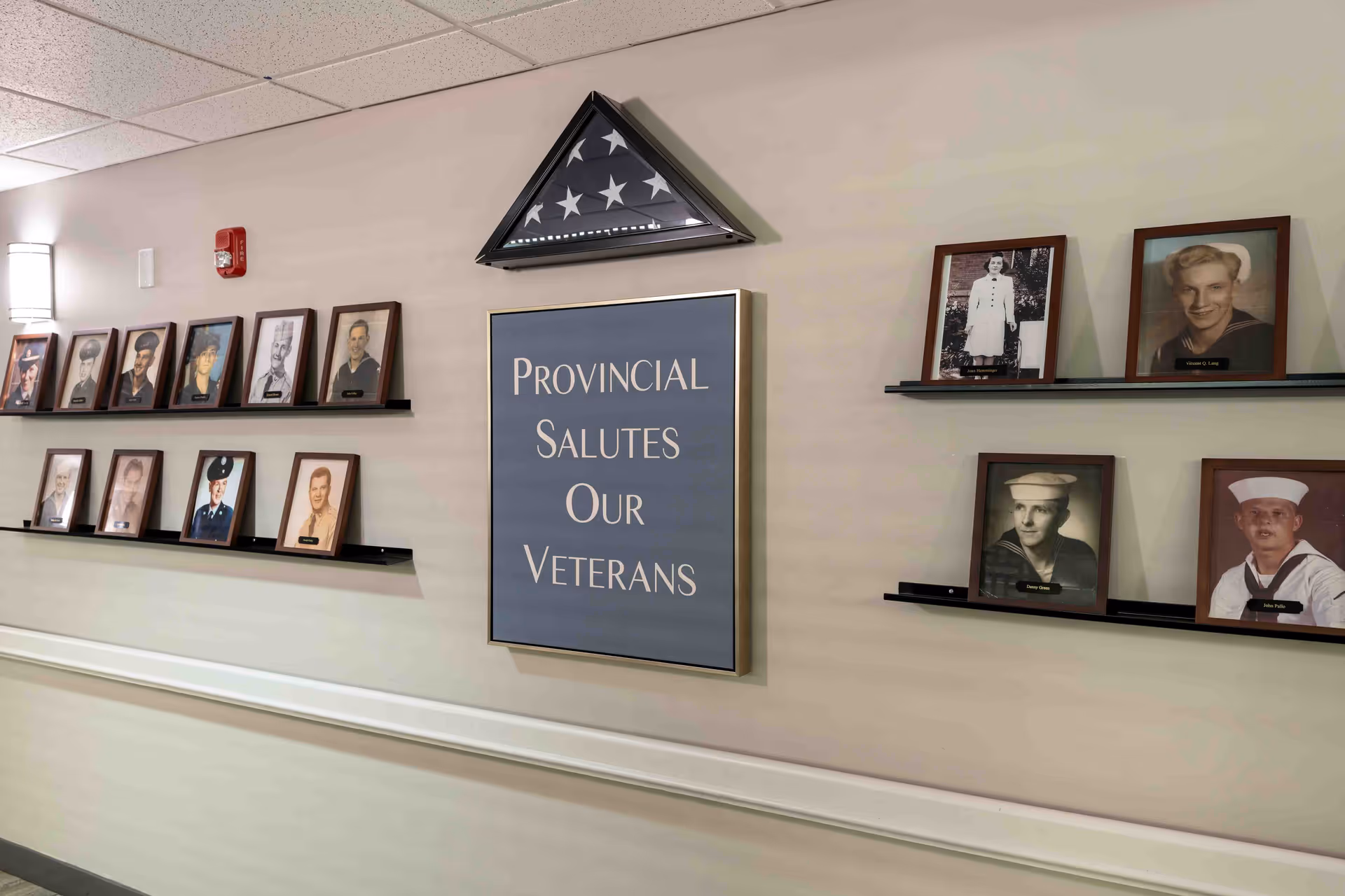 A hallway wall display featuring framed photographs of veterans arranged on black shelves. In the center is a folded American flag in a triangular case above a sign that reads 'Provincial Salutes Our Veterans.' The wall is painted light beige with a white chair rail.
