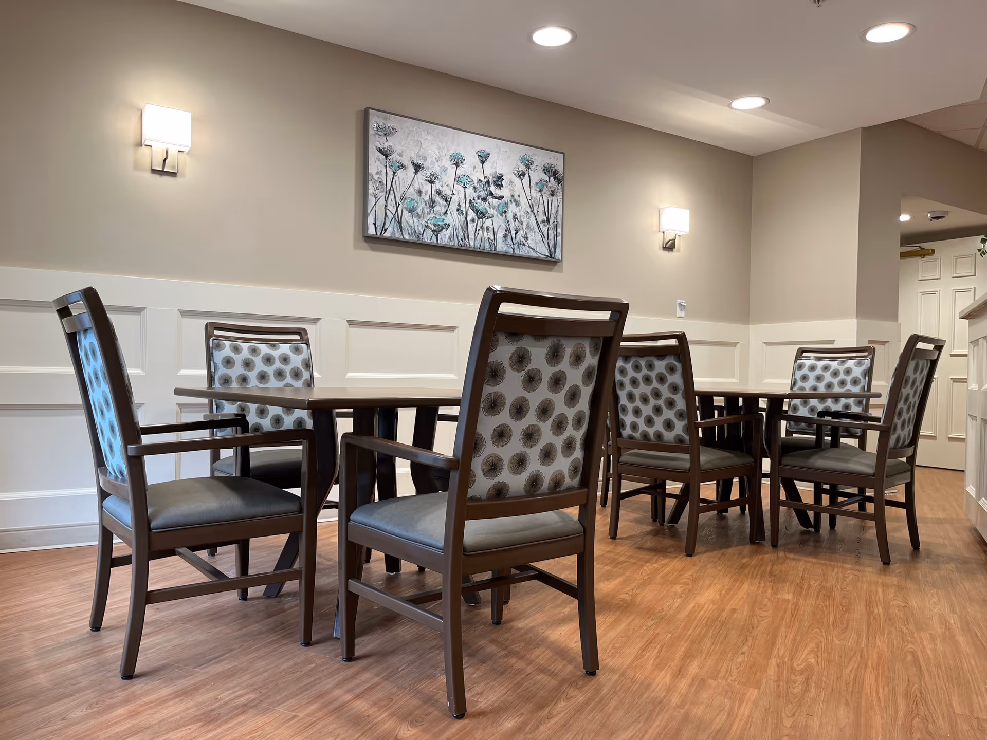 Small dining area with wooden tables and patterned upholstered chairs beneath wall sconces and a floral painting.