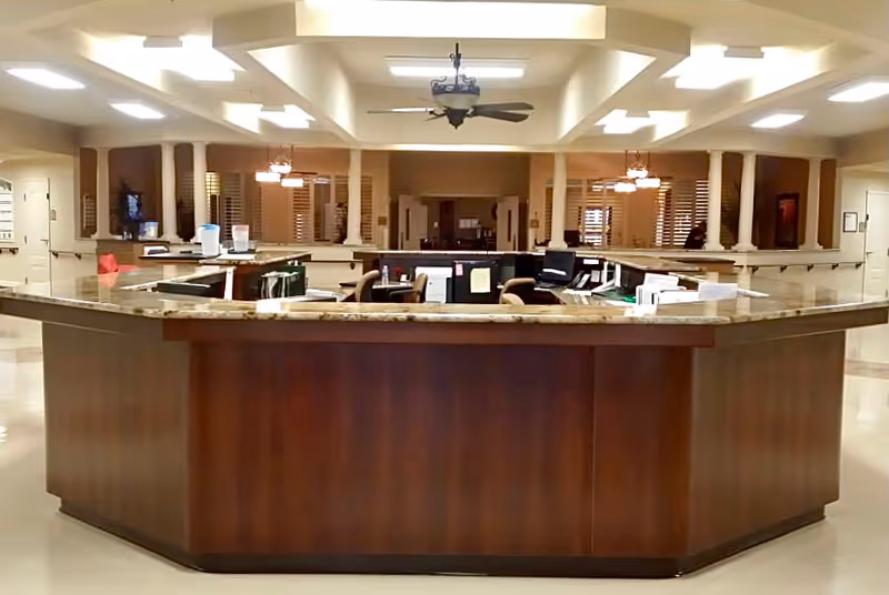 A large reception desk with a polished wooden front and granite countertop in a well-lit interior space. Behind the desk are office chairs, computer monitors, and office supplies. The background features columns, ceiling fans, and warm lighting fixtures, suggesting a welcoming area in a nursing and rehabilitation center.