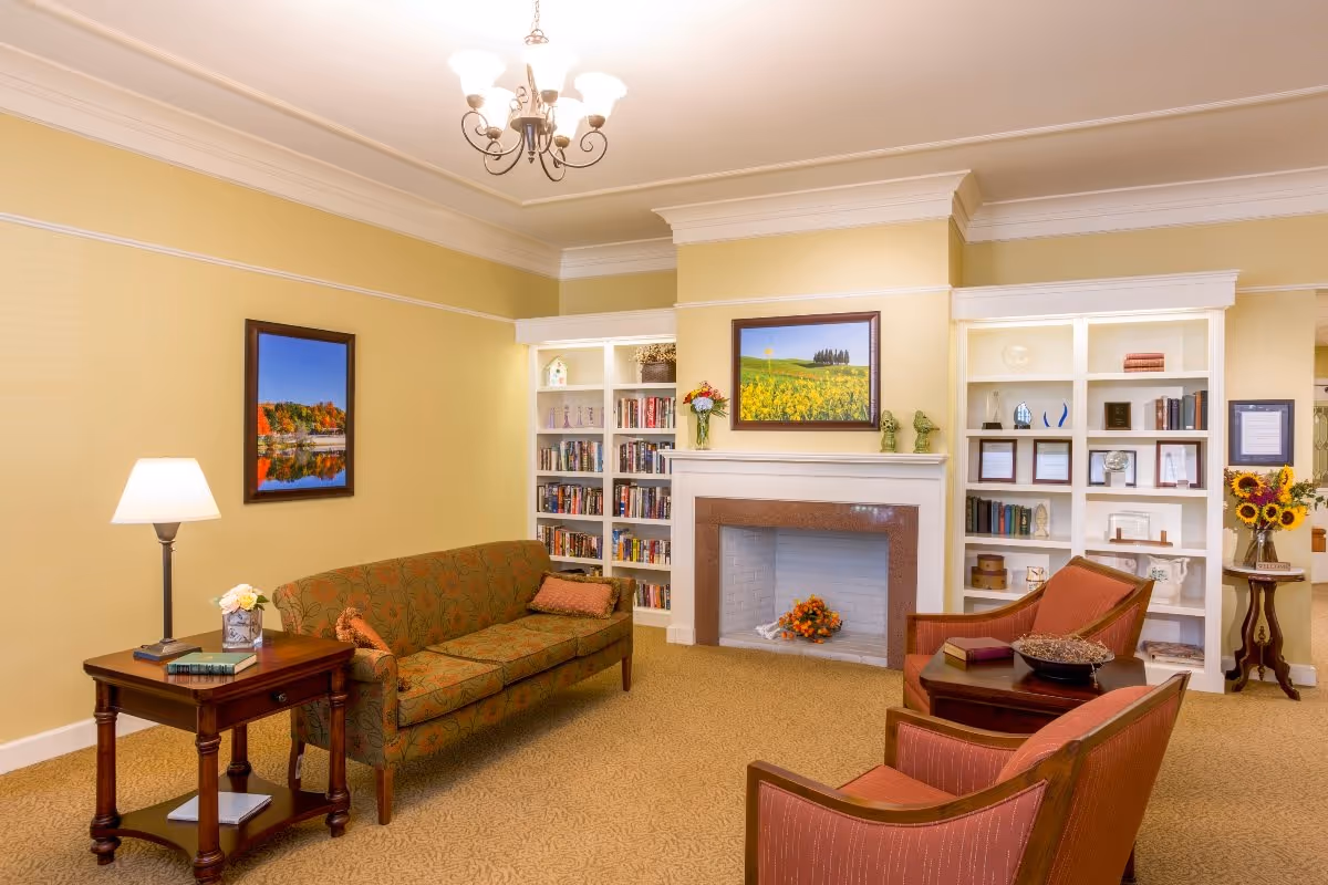 Sunlit living room with a sofa, armchairs, bookshelves and a central fireplace under a chandelier.