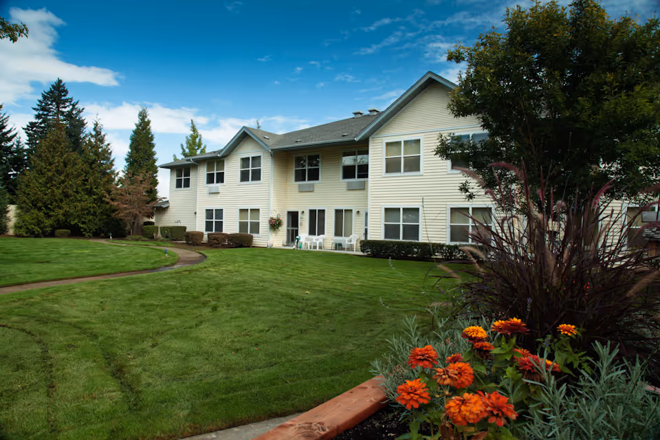 Exterior view of a two-story beige assisted living building with multiple windows, surrounded by a well-maintained green lawn, trees, and colorful flowers in the foreground under a blue sky with some clouds.