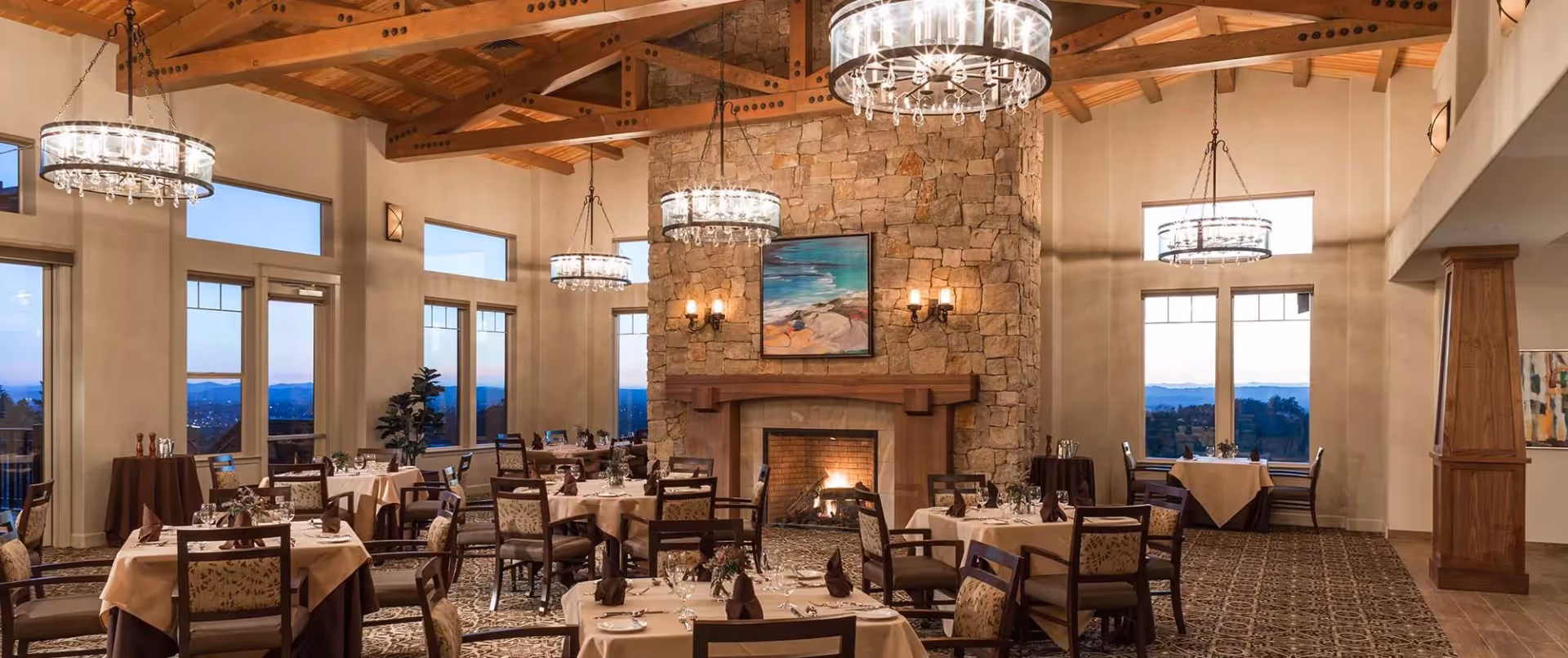 Elegant dining room with multiple tables covered in beige tablecloths and set with glassware and napkins. The room features a large stone fireplace with a painting above it, wooden ceiling beams, large windows with views of the outdoors, and modern chandeliers hanging from the ceiling.