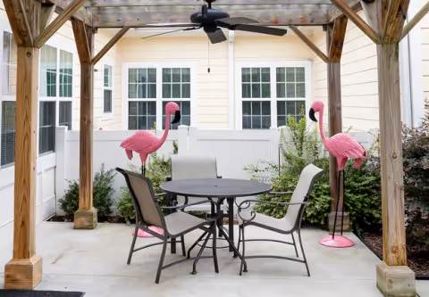 Outdoor patio area with a wooden pergola overhead, a ceiling fan, a round metal table, and four chairs. Two large pink flamingo lawn ornaments stand on either side of the patio, with green bushes and a white fence in the background.
