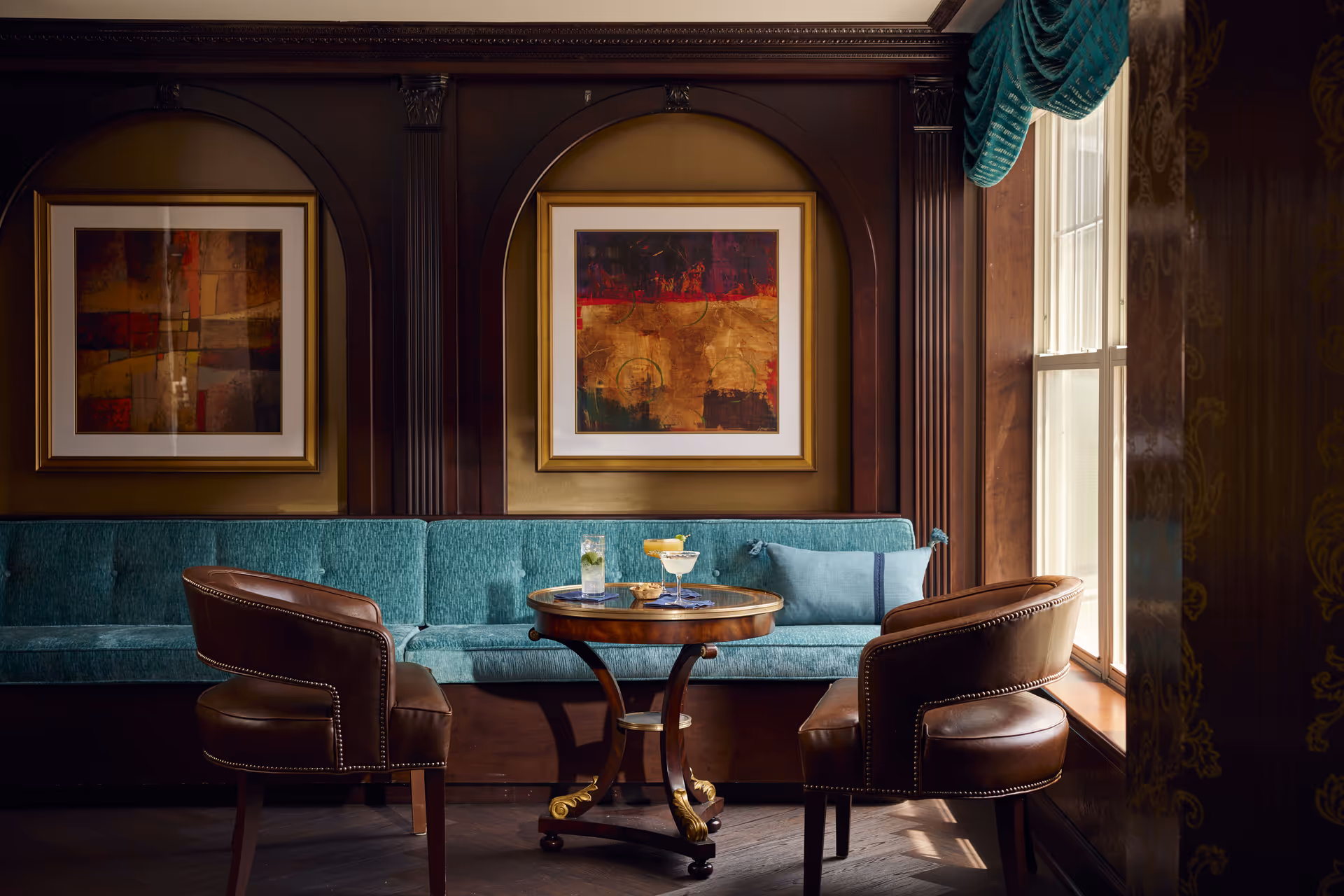 A cozy seating area with a teal upholstered bench along the wall, two brown leather chairs, and a small round wooden table with drinks and a bowl of snacks. The wall behind features dark wood paneling with two framed abstract paintings. A window with teal drapes allows natural light into the room.