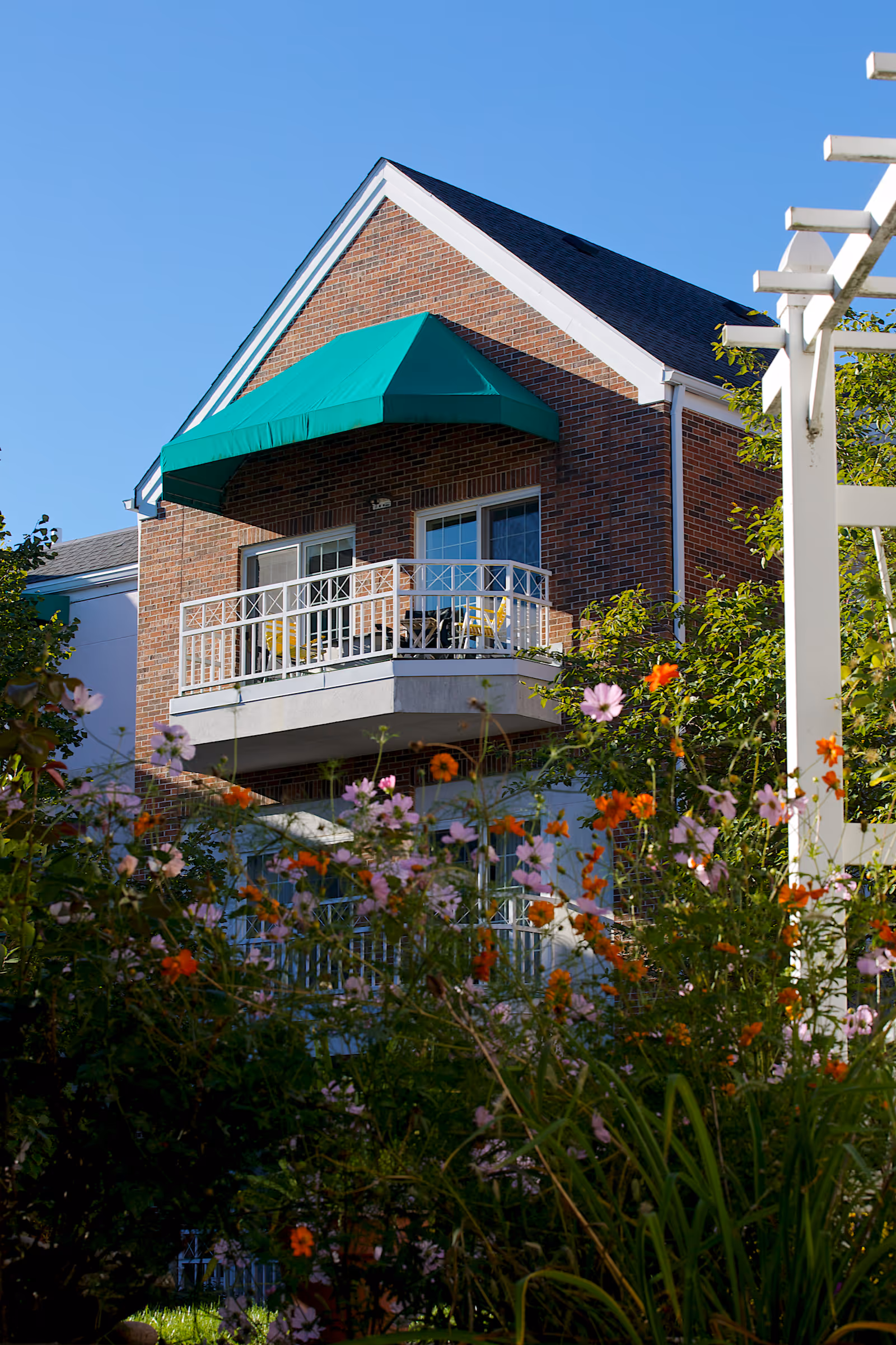 A brick building with a balcony featuring a green awning and white railing, surrounded by colorful flowers and greenery under a clear blue sky.