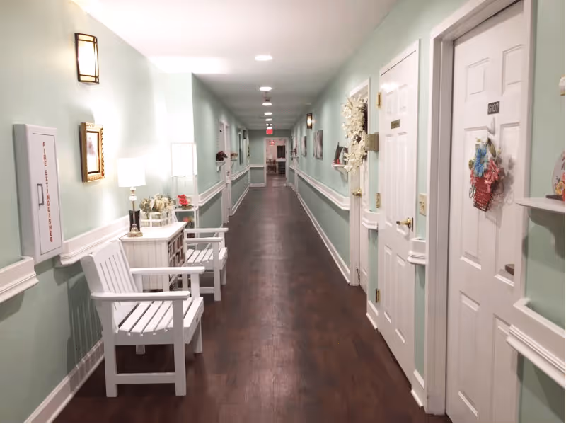 A long, well-lit hallway in an assisted living facility with light green walls, white trim, and dark wood flooring. White wooden chairs and small tables with lamps and decorative items are placed along the left side. Several white doors with room numbers and decorative wreaths line the right side of the hallway. Wall sconces provide additional lighting.