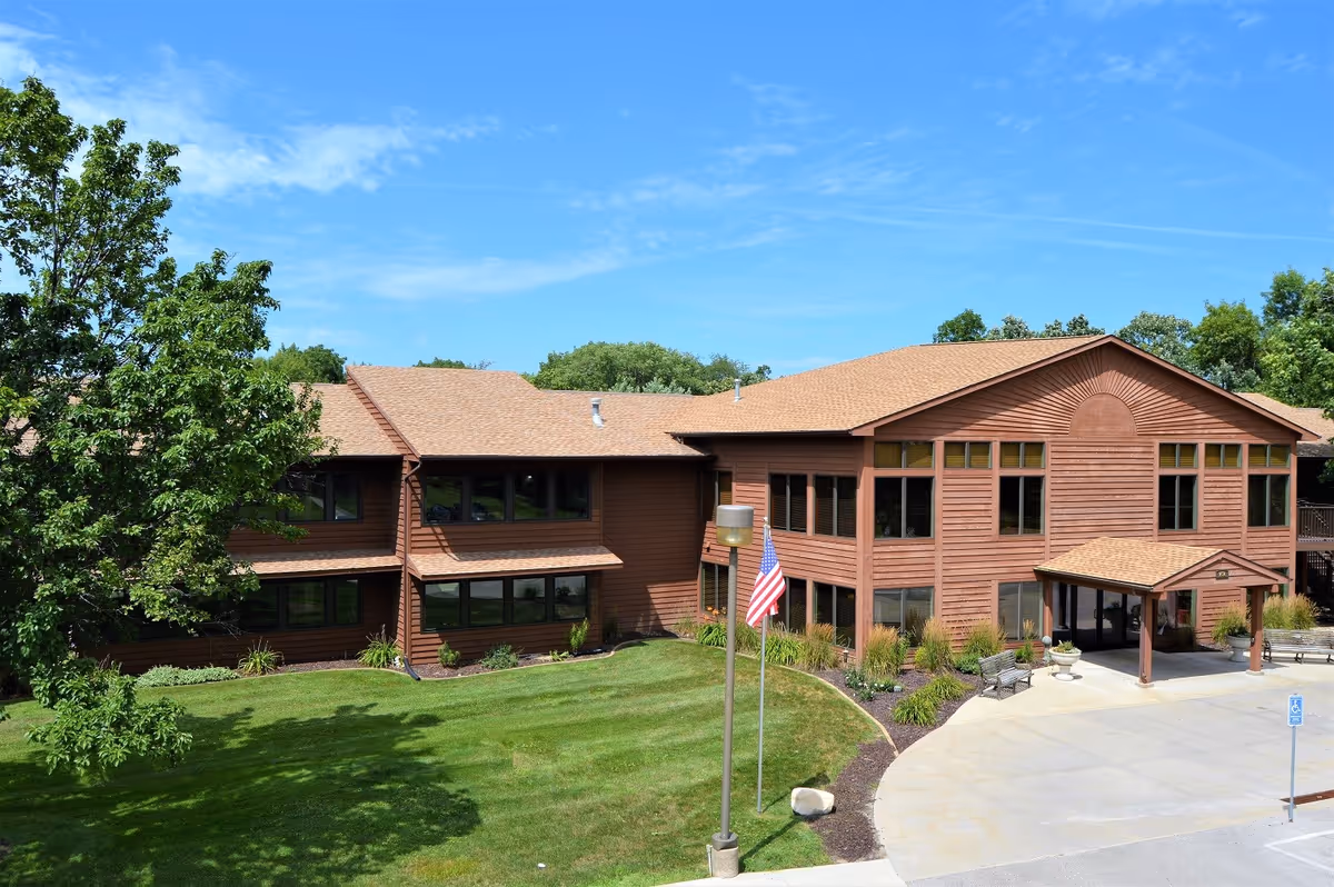 Exterior view of Garnett Place, a two-story building with brown wooden siding and a tan roof, surrounded by green grass, trees, and a clear blue sky. There is an American flag on a pole near the entrance and a paved driveway with a handicap parking sign.
