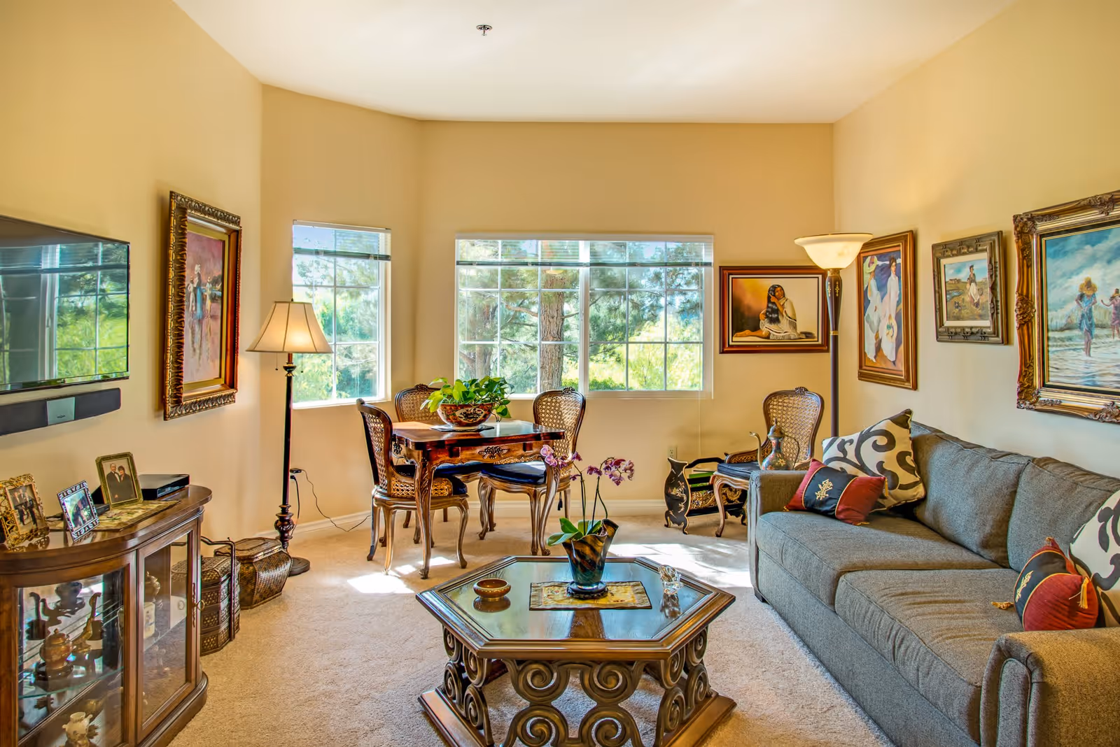 A bright living room with a large gray sofa adorned with patterned and solid pillows, a decorative wooden coffee table with a plant and small items on it, and a small dining table with four chairs near two windows letting in natural light. The walls are decorated with framed paintings, and there are two floor lamps providing additional lighting. A flat-screen TV is mounted on the wall above a wooden cabinet with framed photos and decorative items.