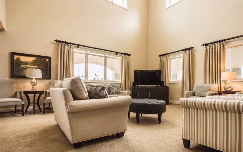 A spacious living room with beige walls and carpet, featuring a beige sofa with patterned cushions, a striped armchair, two additional chairs, a black ottoman, a flat-screen TV on a dark wooden stand, two side tables with lamps, and large windows with beige curtains allowing natural light to fill the room.