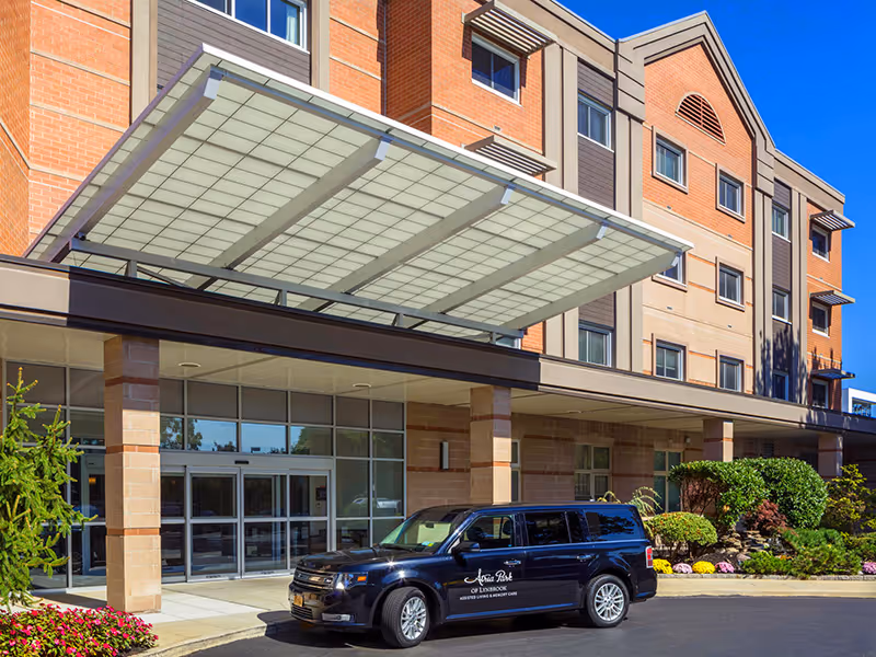 Exterior view of a multi-story senior living facility building with a covered entrance. A black vehicle with the logo 'Atria Park of Lynbrook' is parked in front. The building has large windows and a mix of brick and beige siding with landscaping including bushes and flowers near the entrance.