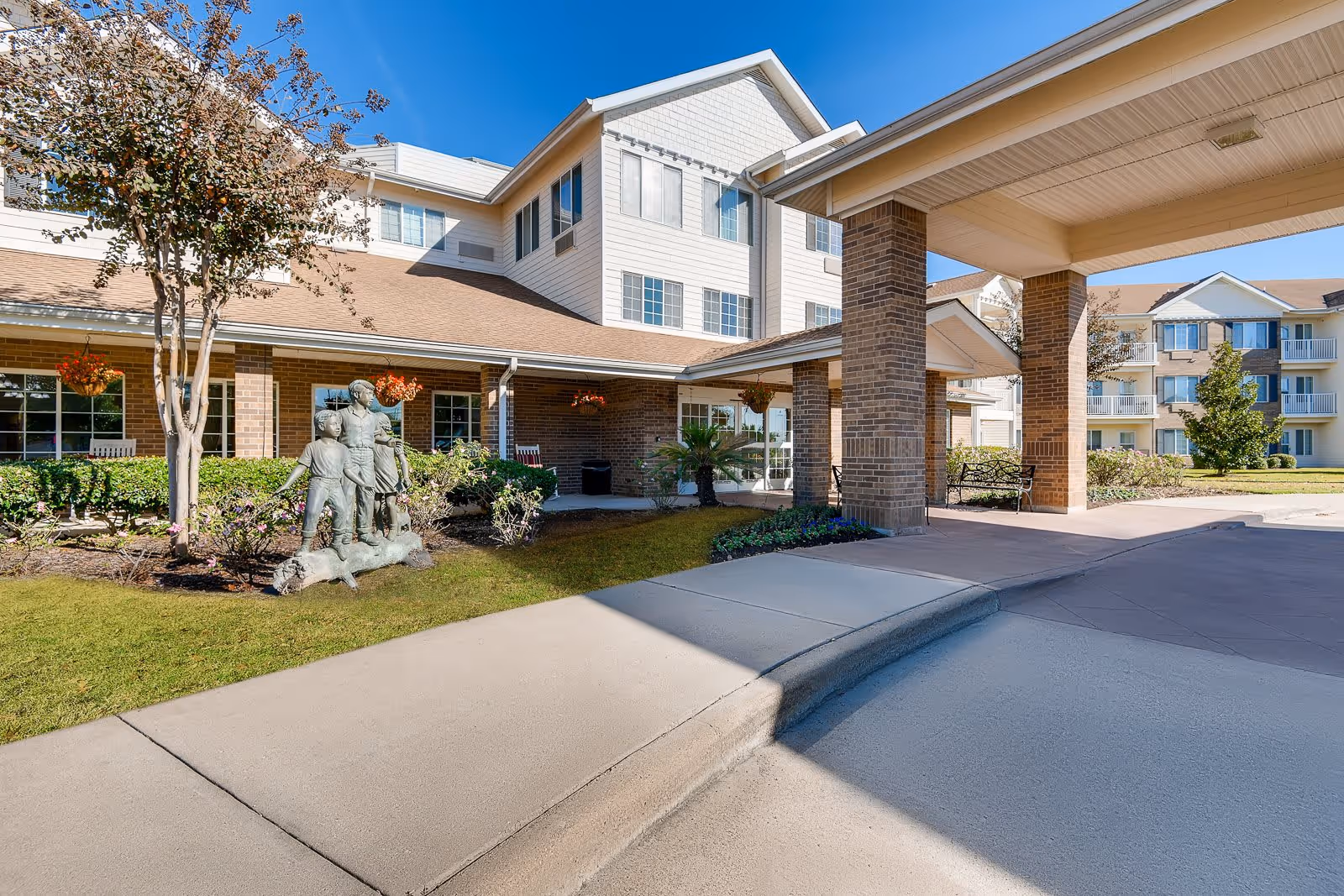 Exterior view of Copperfield Estates - Sky Active Living facility showing a covered entrance with brick pillars, a sidewalk, manicured lawn, and a statue of three children playing near a tree and flower beds under a clear blue sky.