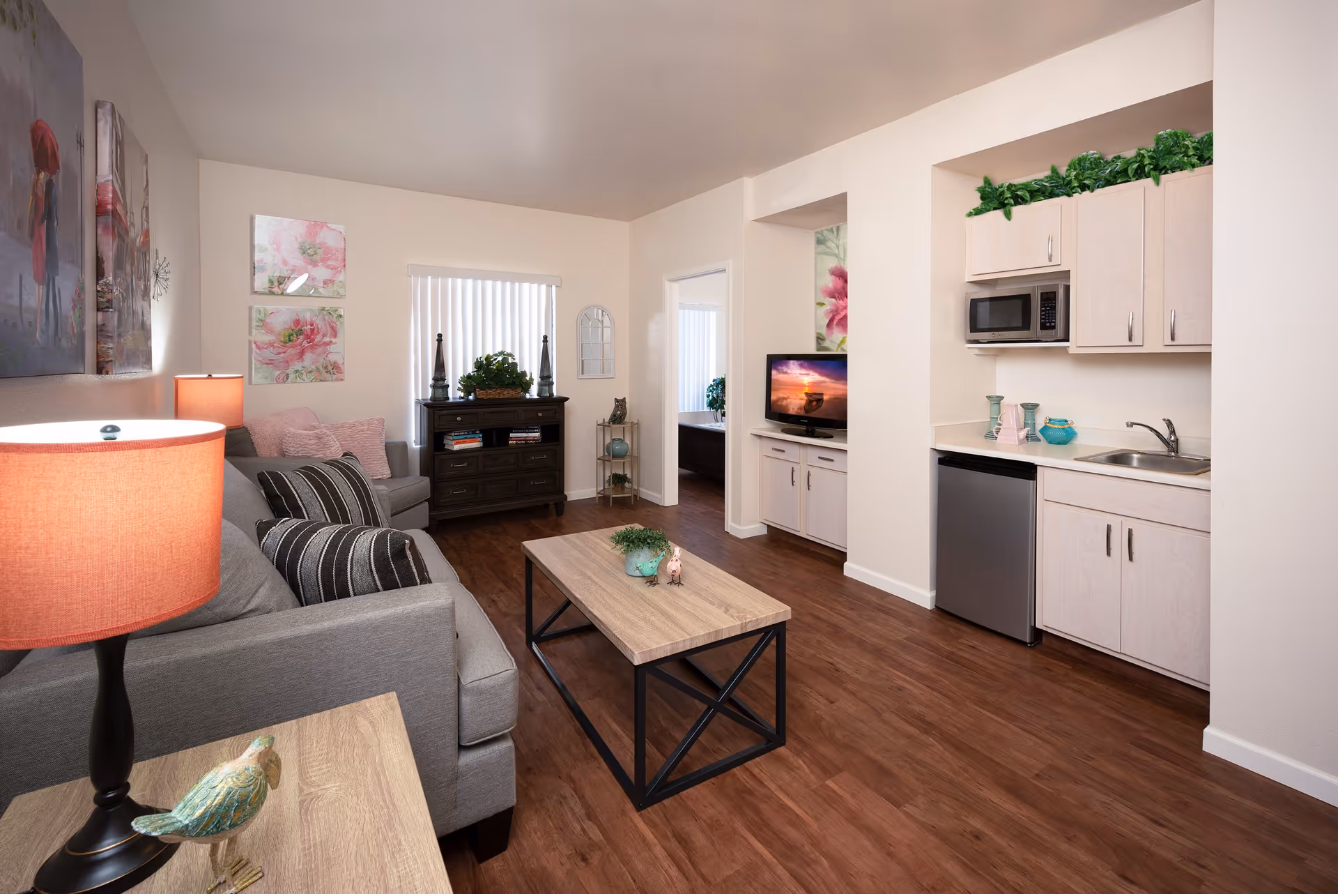 A cozy assisted living apartment living room with a gray sofa adorned with striped and pink pillows, a wooden coffee table with a small plant and decorative figurine, a side table with a lamp and bird figurine, a dark wooden cabinet with plants and books, a flat-screen TV on a white cabinet, and a small kitchenette with light wood cabinets, a microwave, mini fridge, and sink. The room has wood flooring and light-colored walls with floral artwork and a window with vertical blinds.