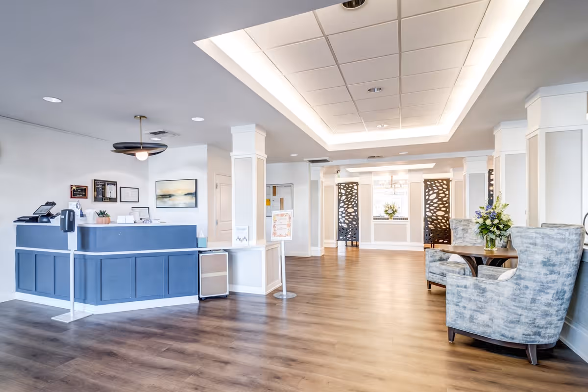 A bright and spacious reception area with a blue front desk on the left side, a modern ceiling light fixture, and a seating area with two upholstered chairs and a small round table with a flower arrangement on the right. The floor is wood, and the walls are white with decorative paneling. In the background, there are decorative black panels and a window with a flower vase on the sill.