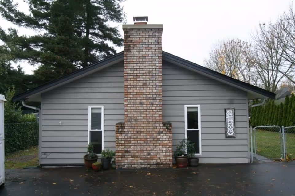 Exterior view of a single-story gray house with a prominent brick chimney in the center, two vertical windows on either side of the chimney, several potted plants near the base of the chimney, a driveway in front, and a fenced yard with trees in the background.