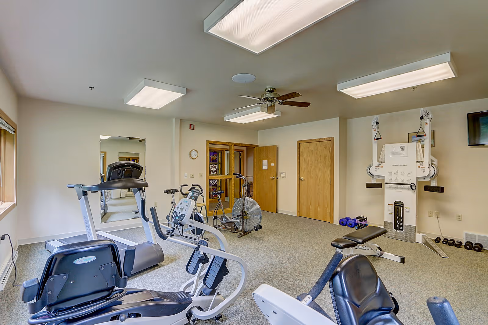 A fitness room with various exercise equipment including a treadmill, stationary bikes, an air bike, a weight bench, and a multi-functional weight machine. The room has beige walls, carpeted floor, ceiling lights, a ceiling fan, a wall clock, and a large mirror on one wall. There are two wooden doors and a window letting in natural light.