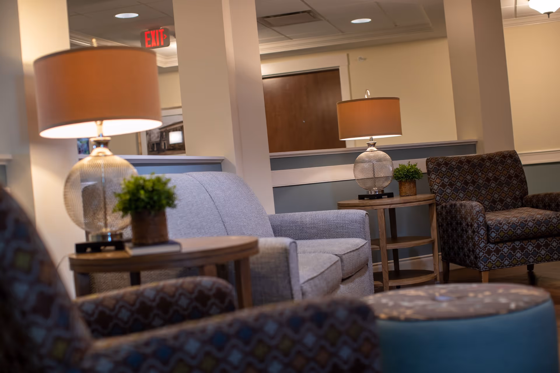 A cozy seating area in a senior living facility with a gray loveseat, two patterned armchairs, wooden side tables with table lamps, and small potted plants. The background shows a hallway with a wooden door and an illuminated exit sign.