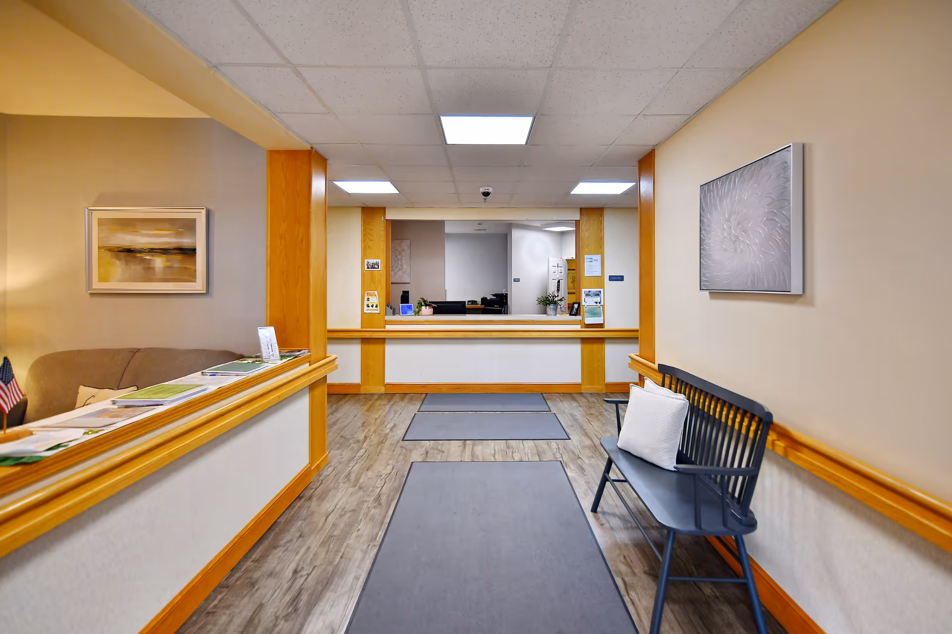 Interior view of a senior living facility reception area with a wooden bench with a white pillow on the right, a counter on the left with brochures and an American flag, two framed artworks on the walls, and a reception desk in the background.