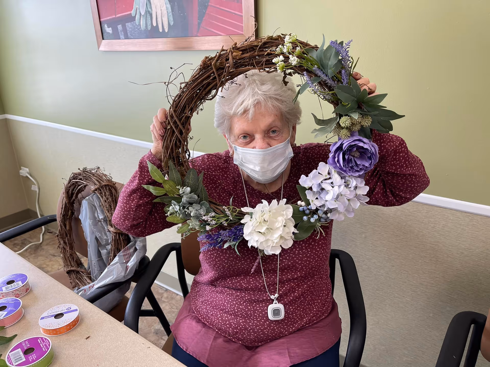 An elderly woman wearing a face mask and a maroon patterned sweater is sitting at a table holding a decorative wreath adorned with artificial flowers and greenery. On the table are several rolls of colorful ribbon. The background shows a green wall with a framed picture and another wreath resting on a chair.