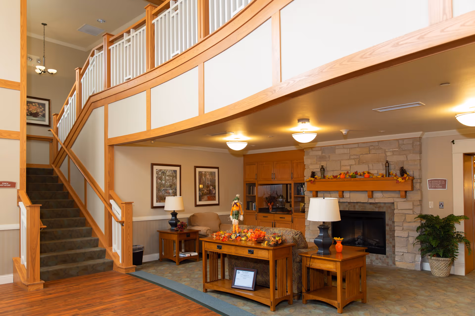 Interior view of a senior living facility lounge area with a stone fireplace decorated with autumn leaves, wooden furniture including tables and lamps, a staircase with wooden railings leading to an upper floor, and framed artwork on the walls.
