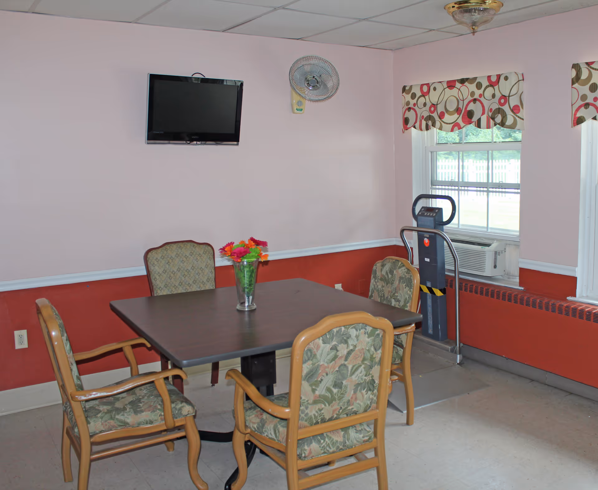 A small room with a square table surrounded by four cushioned chairs with floral upholstery. A vase with colorful flowers is placed on the table. The walls are painted pink with a red lower section and white trim. A flat-screen TV is mounted on the wall above the table. There is a window with patterned valances and an air conditioning unit below it. A standing fan is mounted on the wall near the window.