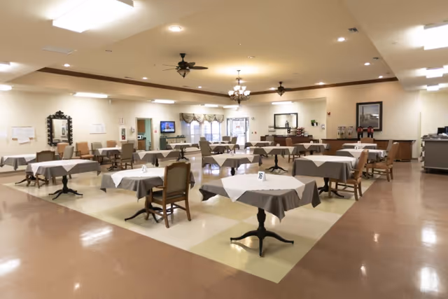 Spacious dining room with multiple tables covered in white and gray tablecloths and chairs arranged under ceiling fans and chandeliers.