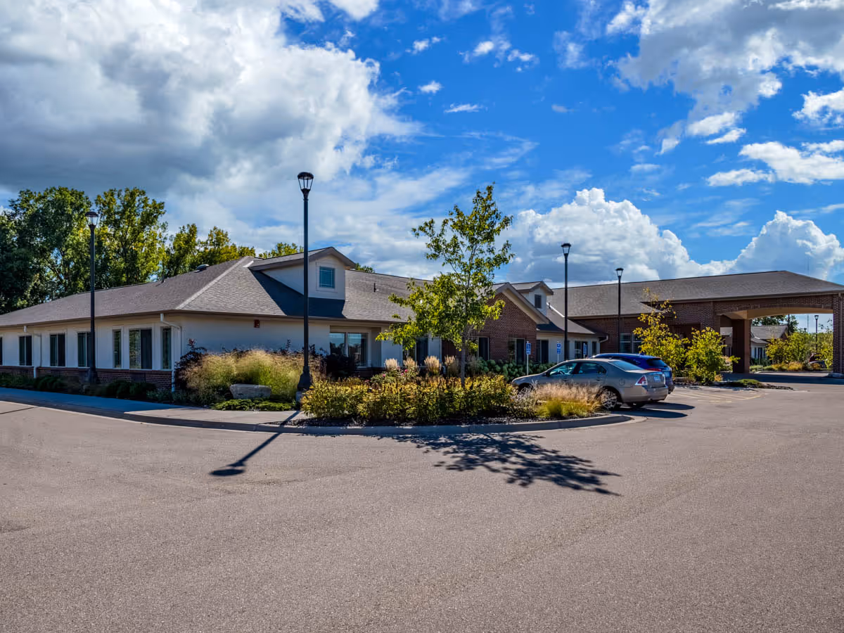 Exterior view of a single-story senior living facility building with a brick and white facade, surrounded by a parking lot with a few parked cars, street lamps, and landscaped greenery under a partly cloudy blue sky.