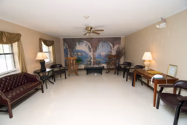 A spacious living room with beige walls and a white tiled floor. The room features a burgundy leather couch on the left, several wooden and black chairs arranged along the walls, two side tables with lamps, and a ceiling fan. The far wall has a mural depicting an outdoor scene with arches, water, and greenery.
