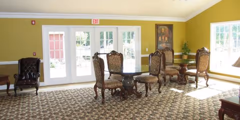 A bright room with patterned carpet, yellow-green walls, and large windows letting in natural light. There are several ornate wooden chairs with upholstered seats and backs arranged around small round tables. A dark leather armchair is positioned near the left wall, and a framed painting hangs on the right wall. Double glass doors lead outside.