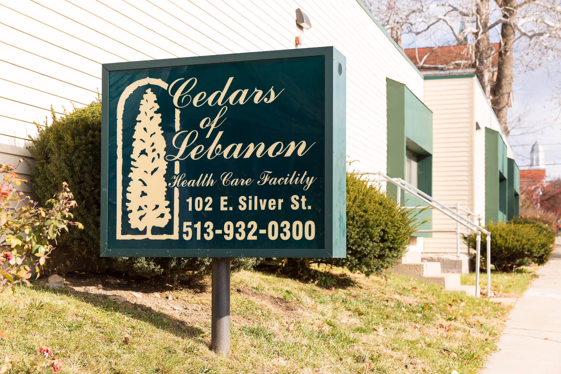 Green sign reading "Cedars of Lebanon Health Care Facility" in front of a light-colored building with shrubs and a sidewalk.