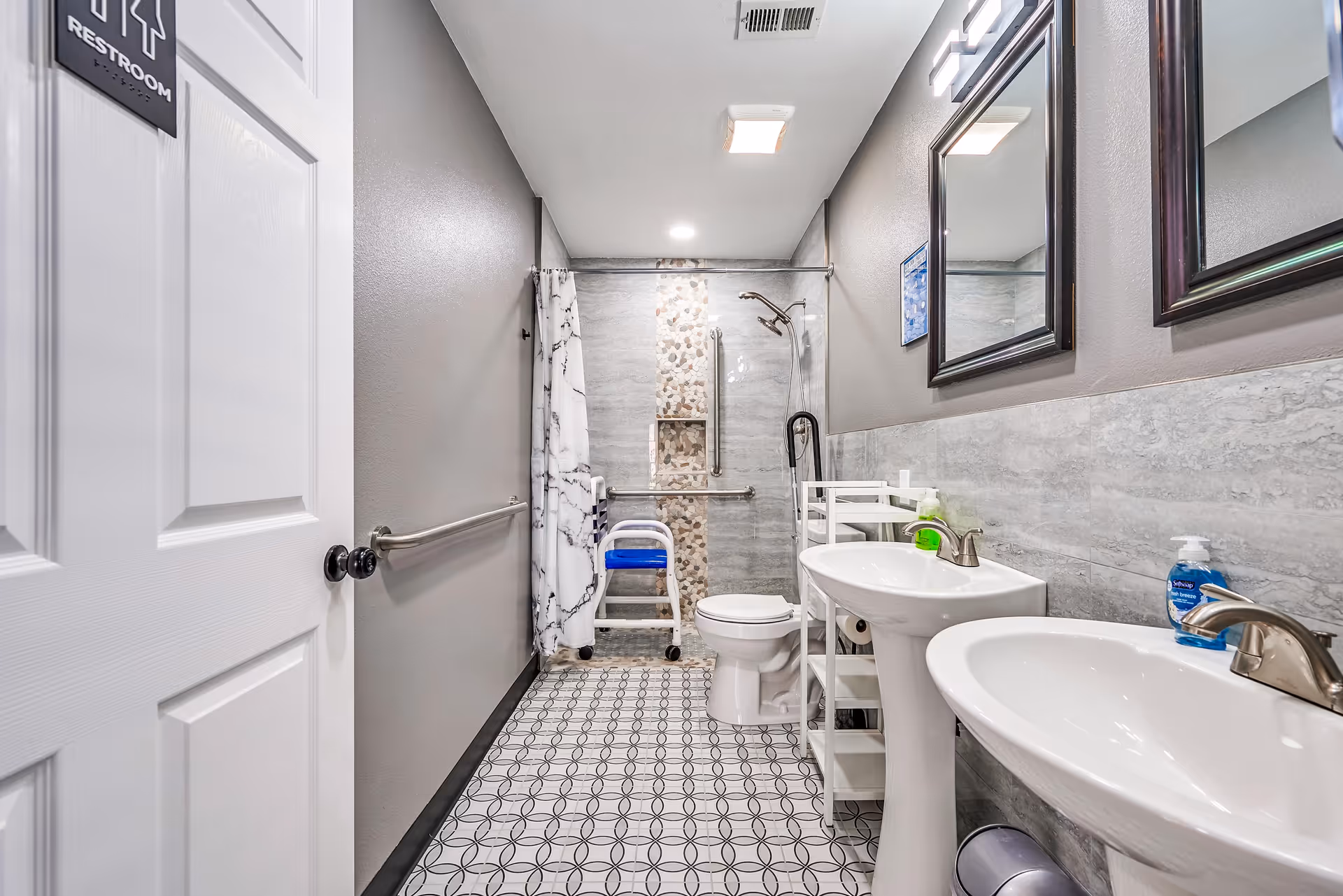 A modern, accessible bathroom with two pedestal sinks, a toilet, and a walk-in shower with a grab bar and a shower chair. The walls are painted gray with a tiled backsplash behind the sinks and shower area. There are two mirrors above the sinks and a patterned tile floor. A restroom sign is visible on the door.