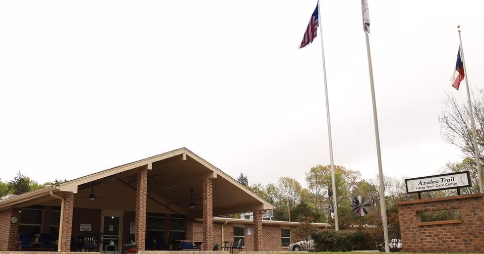 Exterior view of Azalea Trail Long Term Care Center showing a brick building with a covered entrance supported by brick columns. Three flagpoles with flags are in front of the building, along with a sign displaying the facility's name. Trees and shrubs surround the area under an overcast sky.