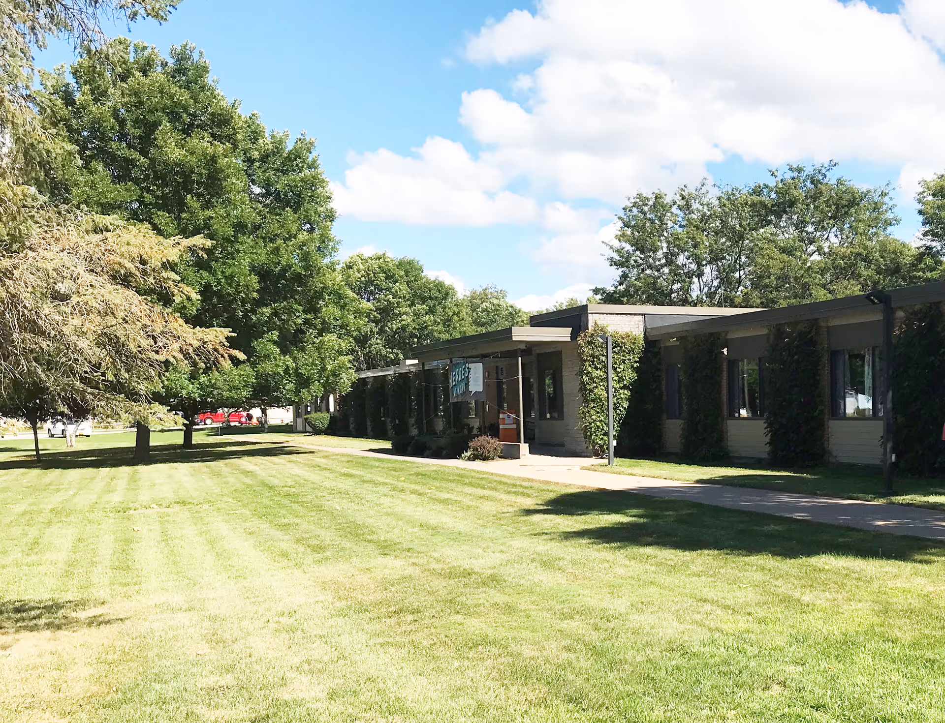 Exterior view of a single-story building with a flat roof, partially covered with ivy, surrounded by a well-maintained lawn and several large trees under a partly cloudy blue sky.