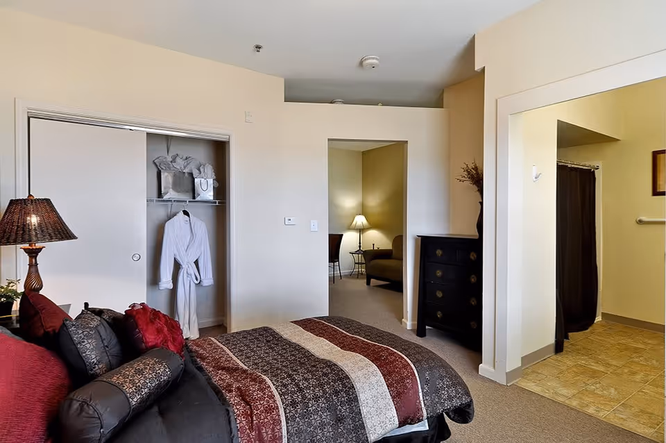 A bedroom in an assisted living facility featuring a bed with a patterned comforter and multiple pillows, a lamp on a side table, an open closet with a white robe hanging inside, a black dresser, and a view into an adjacent sitting area with a lamp and chair. The room also has an open doorway leading to a bathroom with a shower curtain visible.