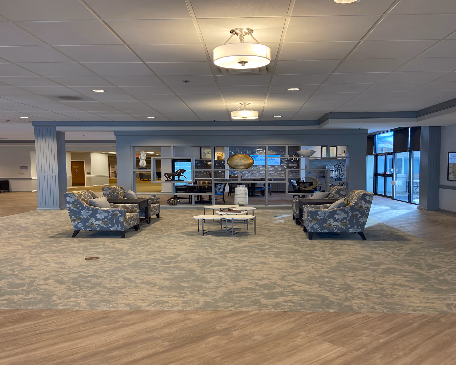 Spacious senior living facility lobby with patterned armchairs arranged around low tables and decorative shelving in the background.