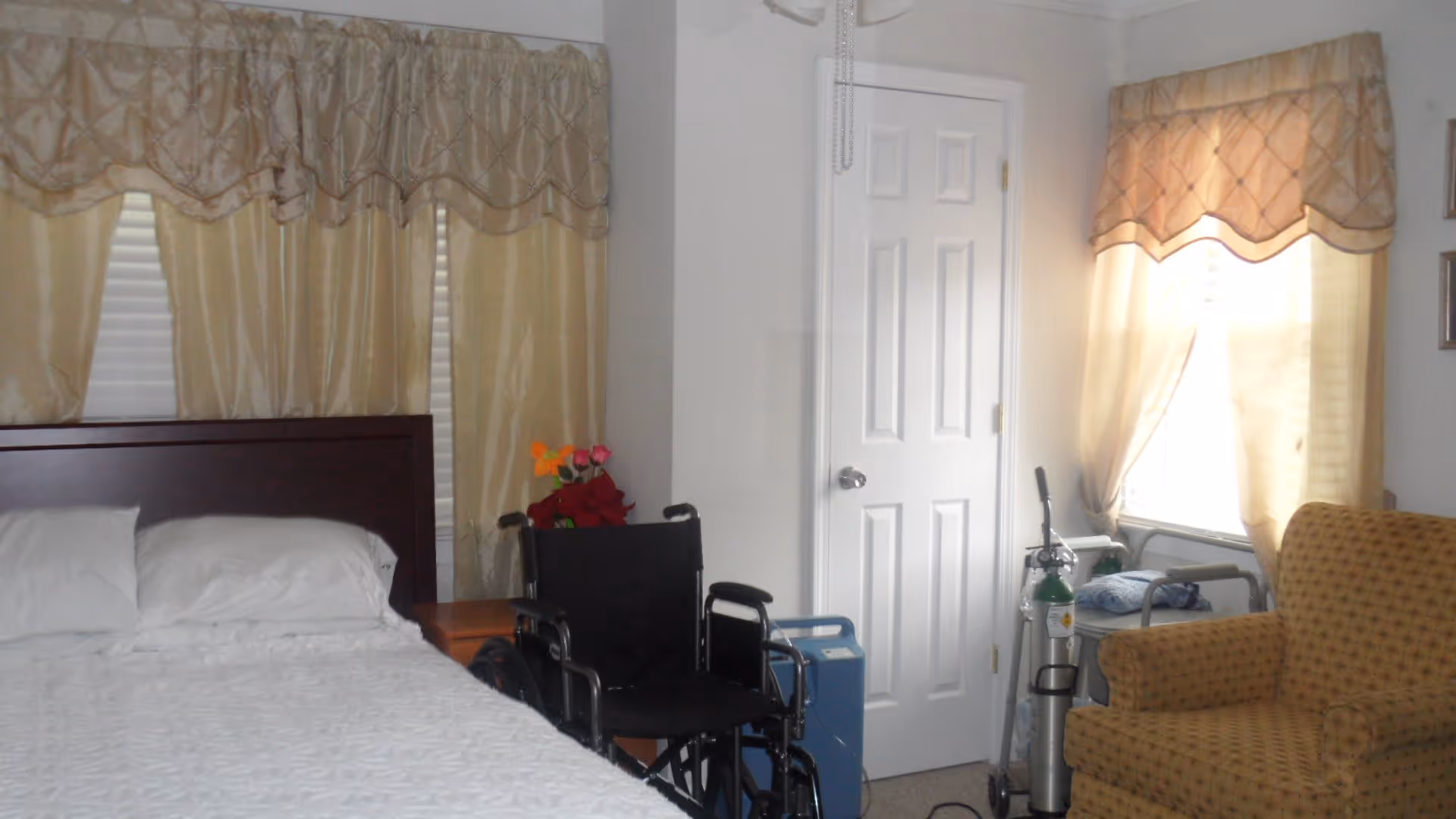 Bedroom with a made bed, a wheelchair and oxygen tank beside a door, and an upholstered armchair by a curtained window.