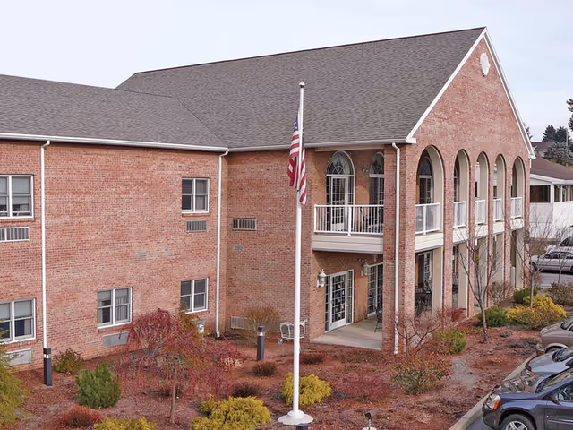 Exterior view of a two-story brick building with a pitched roof, featuring arched windows and a balcony. In front of the building is a flagpole with an American flag, surrounded by landscaped bushes and small trees. Several parked cars are visible on the right side.
