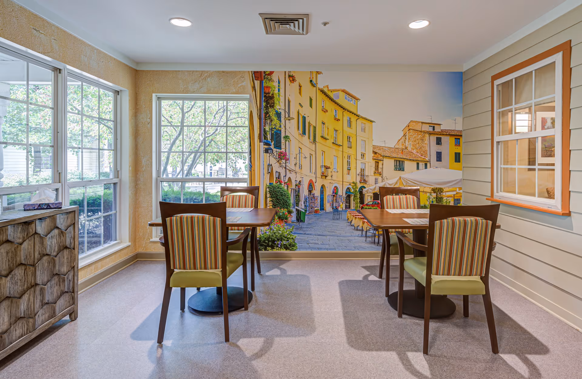 A cozy dining area with two wooden tables and four chairs with striped upholstery. Large windows on the left side let in natural light and show greenery outside. The back wall features a large mural of a European street scene with colorful buildings and outdoor seating. The right wall has a window opening into another room with framed pictures visible inside.