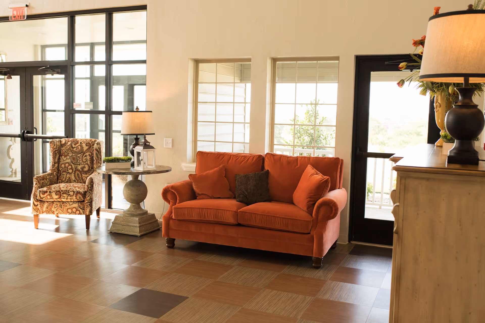 A cozy seating area in a senior living facility with a patterned armchair, a round side table with a lamp and decorative items, and an orange sofa with three cushions. The room has large windows and glass doors letting in natural light, with a wooden floor featuring a checkered pattern.