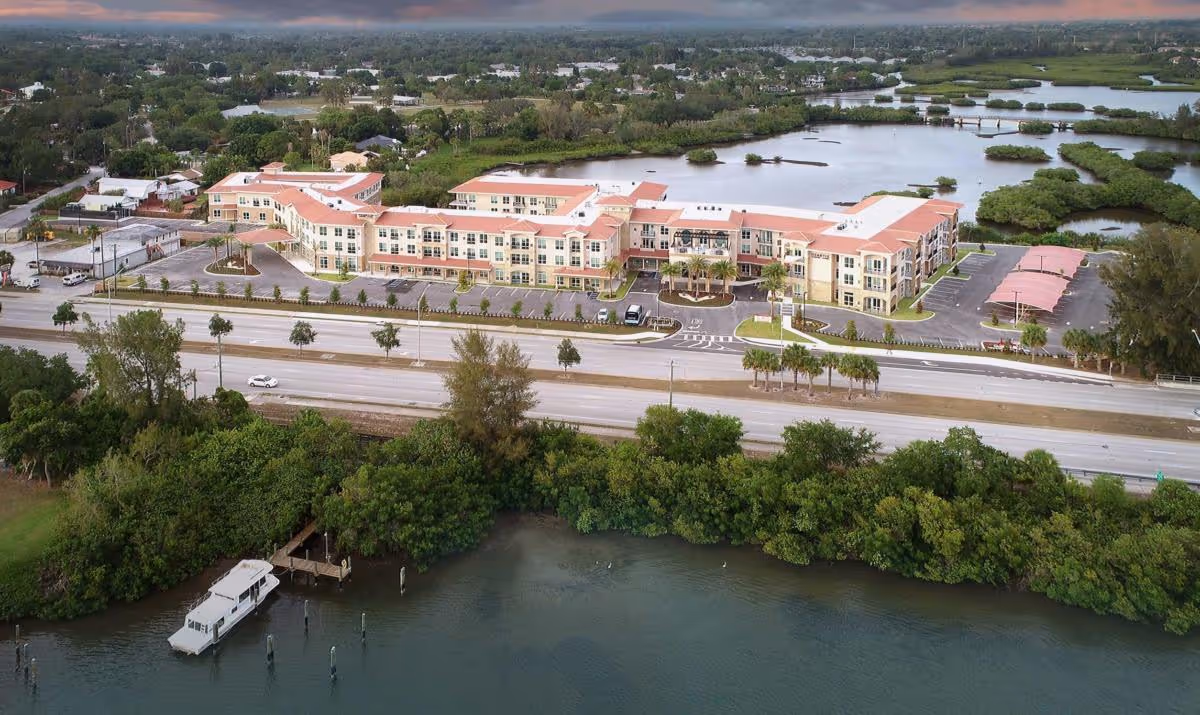 Aerial view of Heartis Venice Assisted Living facility showing a large, multi-story building with a red-tiled roof, surrounded by parking lots, palm trees, and greenery. The facility is located near a body of water with a dock and boat visible in the foreground, and wetlands and residential areas in the background.