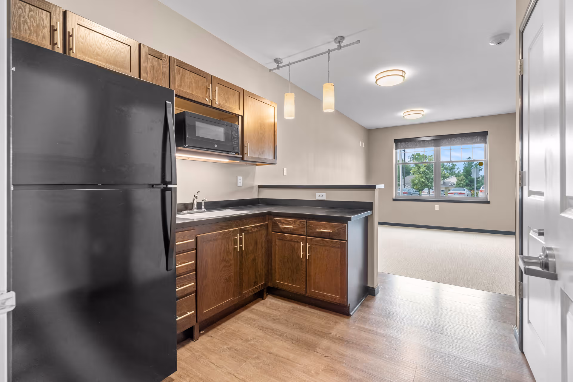 Interior view of a kitchen area with dark wood cabinets, a black refrigerator, a microwave, and a sink. The kitchen opens into a carpeted living space with a large window showing trees and parked cars outside. The room has light beige walls and wood flooring in the kitchen area.