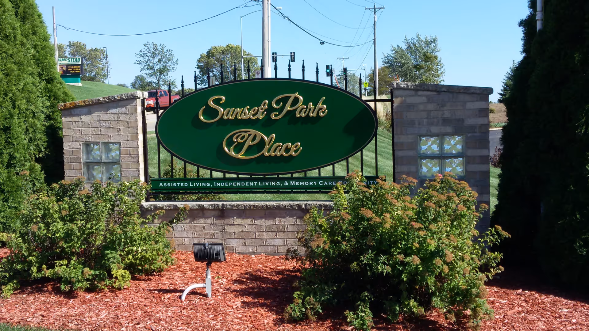 Outdoor entrance sign for Sunset Park Place, a senior living community, surrounded by green bushes and mulch with a clear blue sky in the background.